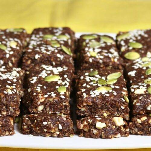 A platter of homemade Dates and Nuts Energy Bars, topped with sesame seeds and pumpkin seeds, arranged neatly on a white plate with a yellow background.