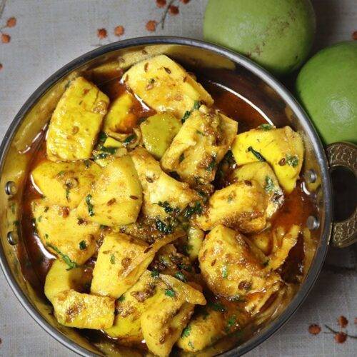 A bowl of vibrant yellow Sweet and Sour Guava Curry, garnished with fresh herbs, with whole guavas placed beside the bowl on a patterned tablecloth.